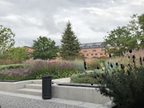A serene garden scene featuring lush greenery and vibrant flowers, with a paved pathway and seating area in the foreground. A large brick building with arched windows towers in the background, surrounded by trees under a cloudy sky.