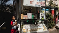 A child holding an ice cream cone stands outside a small kiosk or lottery ticket booth. The booth displays various colorful lottery tickets in the window. There are signs advertising a significant lottery prize amount in Spanish. A person can be seen partially obscured inside the booth, and plants are visible around the area, suggesting an outdoor setting.