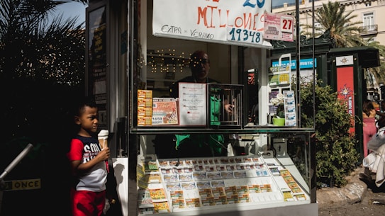 A child holding an ice cream cone stands outside a small kiosk or lottery ticket booth. The booth displays various colorful lottery tickets in the window. There are signs advertising a significant lottery prize amount in Spanish. A person can be seen partially obscured inside the booth, and plants are visible around the area, suggesting an outdoor setting.