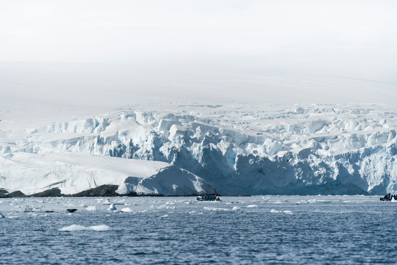 A vast white glacier front rising above the sea, with a tiny inflatable boat beneath it showing the scale