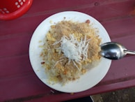 A plate of traditional food featuring sticky rice topped with shredded coconut, ground peanuts, and what appears to be a crumbly mixture of spices or seasonings. A spoon rests on the plate, and the setting is on a reddish-pink table with part of a red basket visible in the corner.