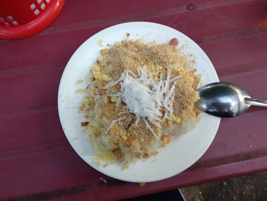 A plate of traditional food featuring sticky rice topped with shredded coconut, ground peanuts, and what appears to be a crumbly mixture of spices or seasonings. A spoon rests on the plate, and the setting is on a reddish-pink table with part of a red basket visible in the corner.