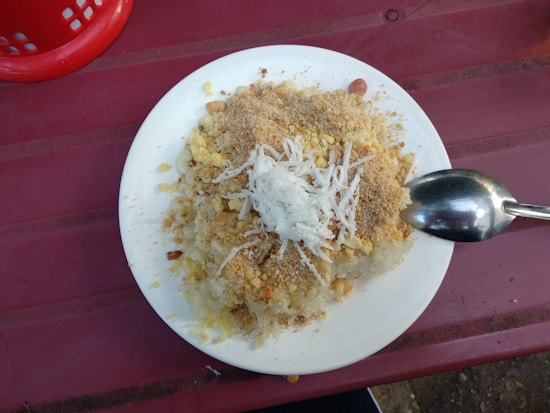 A plate of traditional food featuring sticky rice topped with shredded coconut, ground peanuts, and what appears to be a crumbly mixture of spices or seasonings. A spoon rests on the plate, and the setting is on a reddish-pink table with part of a red basket visible in the corner.