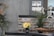A modern kitchen counter features a black granite surface. On the counter, there is a small black vase with bright yellow flowers and a larger black pot with tall green grass-like plants. A decorative wooden bowl sits nearby. The background wall is adorned with rustic brick tiles, and the upper part includes light switches. To the left, there is an arrangement of pink and white flowers.