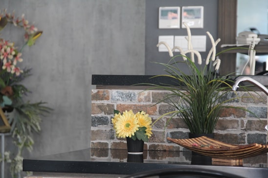 A modern kitchen counter features a black granite surface. On the counter, there is a small black vase with bright yellow flowers and a larger black pot with tall green grass-like plants. A decorative wooden bowl sits nearby. The background wall is adorned with rustic brick tiles, and the upper part includes light switches. To the left, there is an arrangement of pink and white flowers.