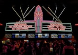 A vibrant, neon-lit concession stand in a theater offering snacks like popcorn, nachos, hot dogs, and ice-cold drinks. People are queued up, visible in silhouette, against the bright menu displays featuring various food items.