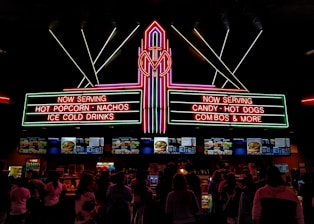 A vibrant, neon-lit concession stand in a theater offering snacks like popcorn, nachos, hot dogs, and ice-cold drinks. People are queued up, visible in silhouette, against the bright menu displays featuring various food items.