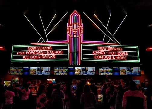 A vibrant, neon-lit concession stand in a theater offering snacks like popcorn, nachos, hot dogs, and ice-cold drinks. People are queued up, visible in silhouette, against the bright menu displays featuring various food items.
