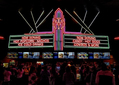 A vibrant, neon-lit concession stand in a theater offering snacks like popcorn, nachos, hot dogs, and ice-cold drinks. People are queued up, visible in silhouette, against the bright menu displays featuring various food items.