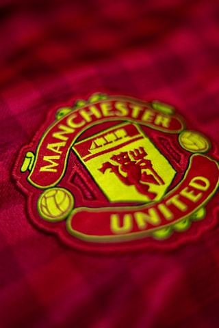 A detailed view of a sports team logo on a red fabric background. The emblem features a shield with a ship, a devil holding a trident, and a football, all set against a yellow and red color scheme. The text surrounding the emblem indicates the name 'Manchester United.'