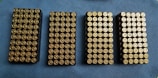 Four rectangular trays filled with neatly arranged brass-colored bullets are placed on a blue textured surface.
