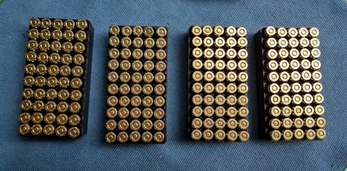 Four rectangular trays filled with neatly arranged brass-colored bullets are placed on a blue textured surface.