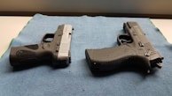 Two handguns are positioned on a blue textured mat, one with a black and silver color scheme and the other entirely black. The surface they are on appears to be well-lit, highlighting the details and textures of the firearms.