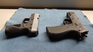 Two handguns are positioned on a blue textured mat, one with a black and silver color scheme and the other entirely black. The surface they are on appears to be well-lit, highlighting the details and textures of the firearms.