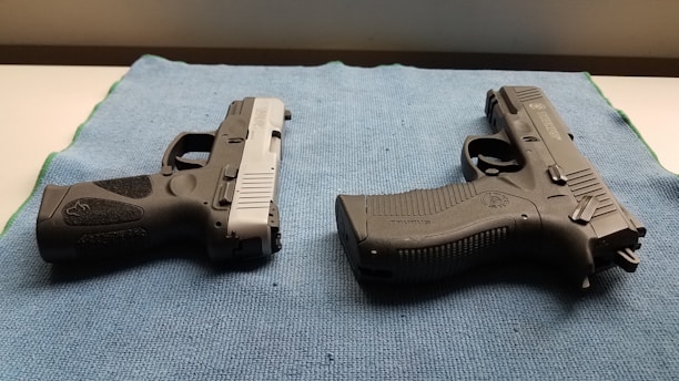 Two handguns are positioned on a blue textured mat, one with a black and silver color scheme and the other entirely black. The surface they are on appears to be well-lit, highlighting the details and textures of the firearms.