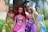 Four dolls dressed as princesses are standing in an outdoor setting with greenery in the background. Each doll has distinct hair color and is wearing a decorative gown in various shades of yellow, pink, purple, and blue.