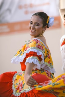 A woman dressed in a vibrant traditional attire is joyfully dancing. Her outfit features bright yellow fabric adorned with floral patterns and lace trim. She has colorful makeup, and her hair is styled up with a decorative hair accessory. Her expression conveys happiness and energy.