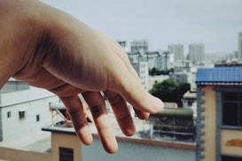 A hand is extended in the foreground against a blurred cityscape in the background. The buildings in the distance appear to be urban residential or commercial structures.