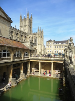 A historic site with a large, green pool surrounded by ancient stone architecture. In the background, a grand cathedral features intricate stonework and tall spires, while people walk and gather around the area. Statues adorn the edges of the structure, and the scene is bathed in natural daylight.
