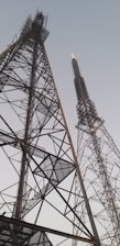 Two tall communication towers with intricate metal frameworks reaching into the sky, set against a pale, clear backdrop.