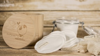 A rustic setting featuring eco-friendly skincare items including cotton pads and a bamboo container. The bamboo container has an illustration and branding. Nearby, there is a clear glass jar with a metal clasp containing a white substance, and cloth pads are spread around a netted bag.