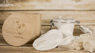 A rustic setting featuring eco-friendly skincare items including cotton pads and a bamboo container. The bamboo container has an illustration and branding. Nearby, there is a clear glass jar with a metal clasp containing a white substance, and cloth pads are spread around a netted bag.