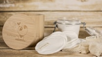 A rustic setting featuring eco-friendly skincare items including cotton pads and a bamboo container. The bamboo container has an illustration and branding. Nearby, there is a clear glass jar with a metal clasp containing a white substance, and cloth pads are spread around a netted bag.