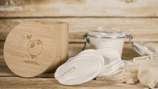 A rustic setting featuring eco-friendly skincare items including cotton pads and a bamboo container. The bamboo container has an illustration and branding. Nearby, there is a clear glass jar with a metal clasp containing a white substance, and cloth pads are spread around a netted bag.