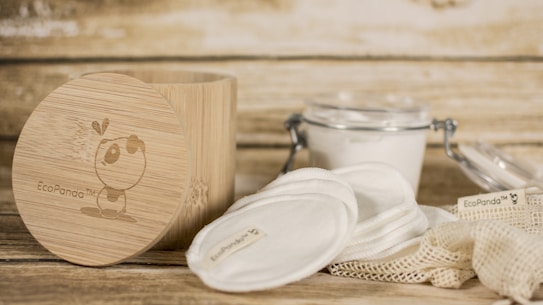 A rustic setting featuring eco-friendly skincare items including cotton pads and a bamboo container. The bamboo container has an illustration and branding. Nearby, there is a clear glass jar with a metal clasp containing a white substance, and cloth pads are spread around a netted bag.