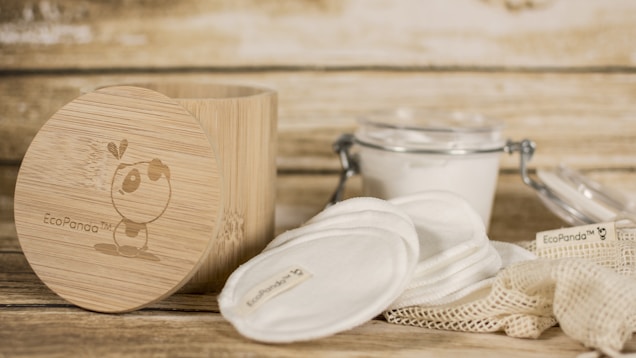 A rustic setting featuring eco-friendly skincare items including cotton pads and a bamboo container. The bamboo container has an illustration and branding. Nearby, there is a clear glass jar with a metal clasp containing a white substance, and cloth pads are spread around a netted bag.