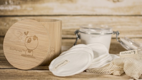 A rustic setting featuring eco-friendly skincare items including cotton pads and a bamboo container. The bamboo container has an illustration and branding. Nearby, there is a clear glass jar with a metal clasp containing a white substance, and cloth pads are spread around a netted bag.