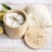On a light wooden surface is a round, bamboo container with a lid beside it. The container holds round white cloth pads tied together with a piece of twine. Next to the container is a netted bag, partially filled with more round white pads. Yellow flowers and green leaves are also present.