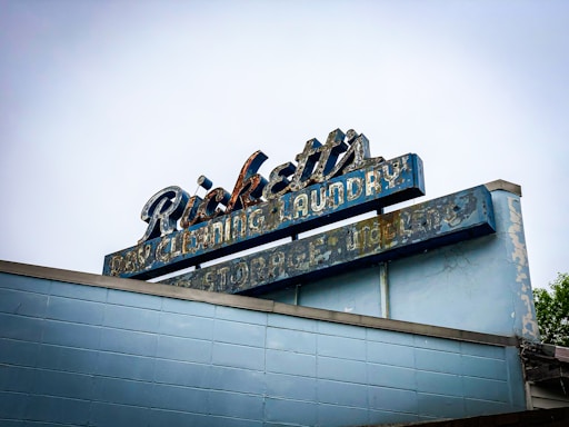 An aged, weathered sign with peeling paint that reads 'Ricketts Dry Cleaning Laundry Storage'. The sign's design features a retro style with faded blue and white colors atop a brick building. The overall condition of the sign suggests it has been exposed to the elements for a long period, giving it a nostalgic, vintage appearance.