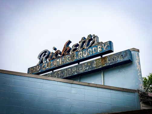 An aged, weathered sign with peeling paint that reads 'Ricketts Dry Cleaning Laundry Storage'. The sign's design features a retro style with faded blue and white colors atop a brick building. The overall condition of the sign suggests it has been exposed to the elements for a long period, giving it a nostalgic, vintage appearance.
