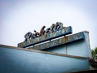 An aged, weathered sign with peeling paint that reads 'Ricketts Dry Cleaning Laundry Storage'. The sign's design features a retro style with faded blue and white colors atop a brick building. The overall condition of the sign suggests it has been exposed to the elements for a long period, giving it a nostalgic, vintage appearance.