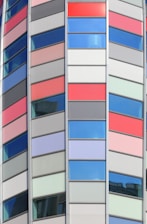 A modern architectural facade featuring a series of vertical panels in red, gray, and blue colors. The windows are interspersed with colorful panels, creating a geometric and vibrant pattern. The design is sleek and contemporary, suggesting a commercial or office building.