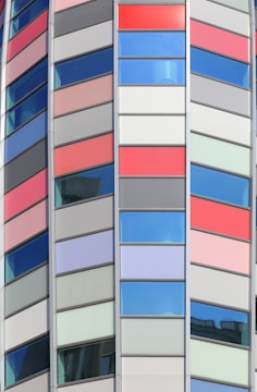 A modern architectural facade featuring a series of vertical panels in red, gray, and blue colors. The windows are interspersed with colorful panels, creating a geometric and vibrant pattern. The design is sleek and contemporary, suggesting a commercial or office building.
