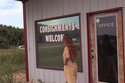 A woman in a yellow checkered dress stands in front of a building with a large window displaying the words 'CONSIGNMENTS WELCOME' in bold letters. To her right is a door with the word 'OFFICE' on it. The surroundings include grasses and trees, and a vehicle is partially visible. The building has a rustic appearance with a corrugated metal exterior.