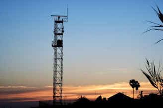 A tall metal tower with a radar on top is silhouetted against a colorful sunset sky. The sky transitions from a deep blue at the top to soft oranges and pinks near the horizon. Palm trees and other vegetation are visible in the foreground, while a small building can be seen to the right of the tower.
