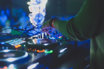 A DJ is mixing music on a console, surrounded by various controls and illuminated by colorful lights. The hand of the DJ is seen in focus, adjusting knobs, implying a lively and energetic nightlife scene.