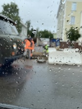 Raindrops cover a window, blurring the view of a street scene outside. In the background, construction workers in high-visibility clothing are near a vehicle and some large concrete barriers. Trees and multi-story buildings are visible under an overcast sky.