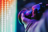 A close-up view of a person holding a professional camera with a large lens, capturing a colorful display of vibrant lights in the background. The scene is illuminated by a spectrum of neon colors ranging from red and orange to green and blue.