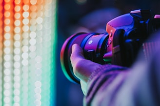 A close-up view of a person holding a professional camera with a large lens, capturing a colorful display of vibrant lights in the background. The scene is illuminated by a spectrum of neon colors ranging from red and orange to green and blue.