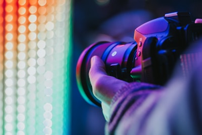 A close-up view of a person holding a professional camera with a large lens, capturing a colorful display of vibrant lights in the background. The scene is illuminated by a spectrum of neon colors ranging from red and orange to green and blue.