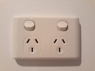 An electrical wall socket with two outlets and individual power switches, set against a light-colored wall.