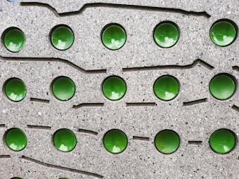 A textured gray surface features a grid pattern of shiny, reflective green circular elements embedded within it. The surface has a speckled, stone-like appearance with grooves and lines surrounding the circular elements.