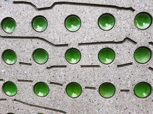 A textured gray surface features a grid pattern of shiny, reflective green circular elements embedded within it. The surface has a speckled, stone-like appearance with grooves and lines surrounding the circular elements.