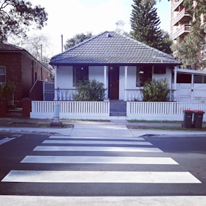 A small, quaint house with a tiled roof and a white picket fence. The front porch features columns and there is lush greenery on either side of the entrance. A zebra crossing is visible in the foreground, leading from the street to the house's entrance. Nearby buildings and trees are visible in the background.