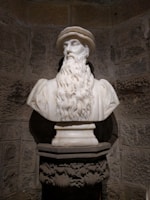 A marble bust of a bearded man wearing a hat is placed on an ornate pedestal against a rustic stone wall, showcasing intricate details and lifelike craftsmanship.