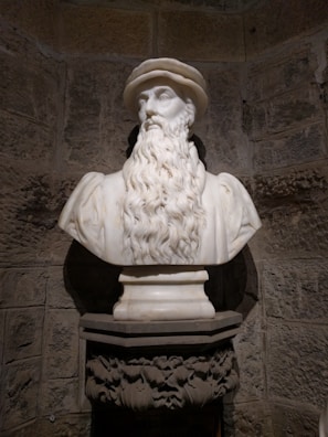 A marble bust of a bearded man wearing a hat is placed on an ornate pedestal against a rustic stone wall, showcasing intricate details and lifelike craftsmanship.
